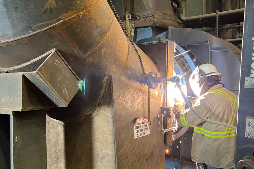 Welder in protective gear performing on-site industrial repair on large metal equipment.