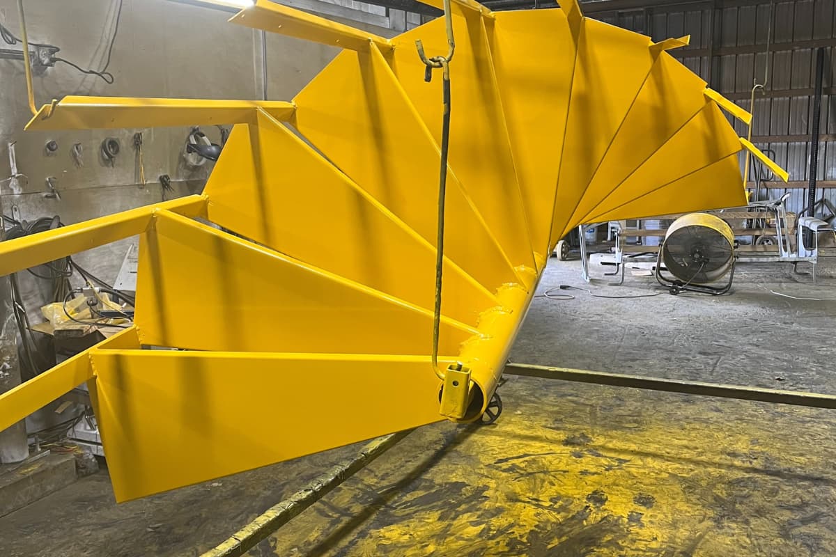 Bright yellow metal part suspended for powder coating inside the WiseCo shop.